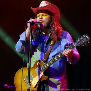 Male musician wearing a cowboy hat sings into a microphone while playing a sunburst Les Paul electric guitar on stage under colorful lights.