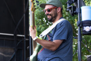 Man in a cap and orange sunglasses plays a bass guitar on an outdoor stage.