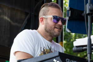 Musician wearing white T-shirt and purple sunglasses plays a keyboard on an outdoor stage.