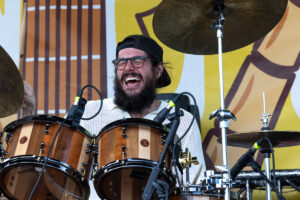 Bearded drummer wearing a backward cap and glasses, mid-performance behind wooden drums on a colorful stage backdrop.