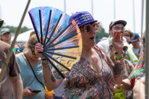 Woman at an outdoor festival waves a large colorful fan, wearing a purple cap and glittery sunglasses amid a cheering crowd.