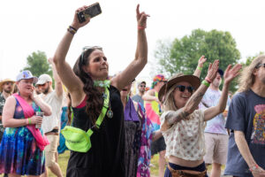Woman at an outdoor festival raises arms and snaps a selfie with her phone, wearing a neon green fanny pack.