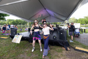 Two women dance and pose for the camera on a park festival stage under a large tent, with a guitarist and photographer in the background.