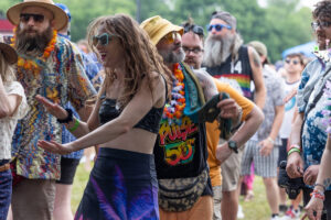 Group of festival-goers dancing outdoors, wearing colorful outfits, leis, and sunglasses in a sunny park setting.