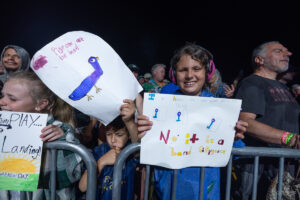 Smiling child with pink headphones holding a handmade poster at a night-time crowd behind a barrier.