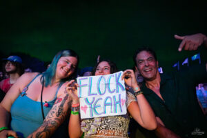 Three concertgoers smile and hold a handmade sign that reads 'FLOCK YEAH'.