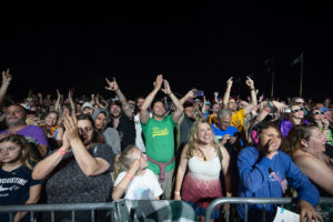 Crowd at a nighttime concert, people clapping and smiling behind a metal barrier in front of the stage.
