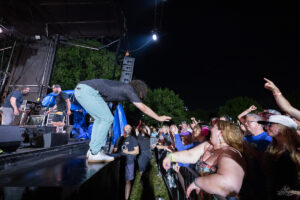 Musician leans offstage toward a cheering crowd at night, reaching hand to fans near the barrier.
