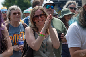 Smiling woman in sunglasses claps her hands at an outdoor festival crowd.