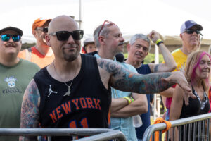 Tattooed, bald man wearing a black New York jersey and sunglasses gestures over a barricade at an outdoor crowd event behind him.