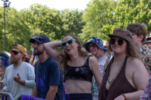 Festival crowd enjoying a performance; woman in blue sunglasses smiles while leaning back, surrounded by friends in hats and colorful outfits.