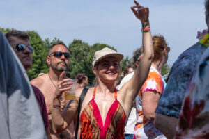 Smiling woman in a colorful dress raises her right arm at a sunny outdoor festival, holding a plastic cup of beer.