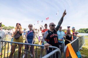 Crowd of diverse festival-goers behind a metal barrier, a tattooed man in a Knicks jersey points upward near the lake.