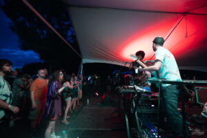 Band performs on stage under a white canopy at night, red lighting and a cheering crowd in front of them.