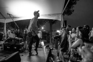 Black-and-white photo of a guitarist on stage under a tent, performing to a crowd at night.