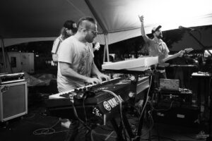 Black-and-white photo of a keyboard player in the foreground performing on a tented stage, with a guitarist singing or speaking into a mic in the background.