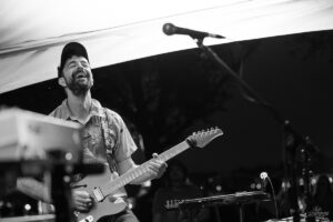 Male guitarist smiling and playing an electric guitar on a stage under a white canopy, microphone overhead nearby