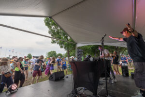Outdoor music festival stage under a white tent with a DJ and a performer reaching toward the crowd; audience dancing and cheering in front of the stage.