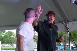 Two men on an outdoor stage under a tent high-fiving, one in a white T‑shirt and sunglasses, the other in a black shirt with headphones around his neck.