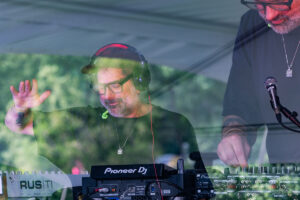 Bearded DJ with glasses and headphones raises hand while mixing on Pioneer DJ gear under a tent, outdoor scene behind glass.