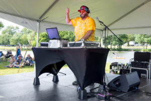 DJ in a bright yellow tie-dye shirt with red sunglasses and headphones, mixing at an outdoor stage under a canopy with equipment nearby.