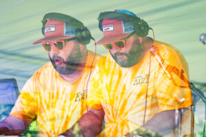 Two bearded DJs in matching yellow tie-dye shirts and orange sunglasses perform side by side on stage, wearing headsets and hats.