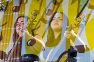 Two women play bass guitars on stage, one with eyes closed, amid a bright yellow abstract backdrop and overlapping reflections for a layered performance look.