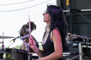 Drummer with purple hair smiling on stage, holding a pink drumstick over a black drum set with gear in the background