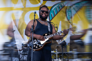 Smiling man in a black tank top and sunglasses plays a black Les Paul guitar on a bright yellow stage backdrop.