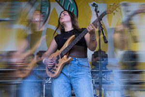 Female bassist on stage playing a natural wood bass guitar, wearing a black top and jeans, with microphones nearby and a yellow backdrop.