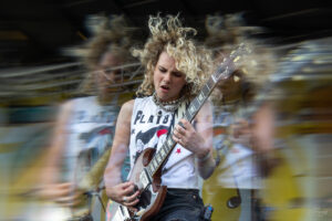 Female guitarist with curly blond hair playing a brown electric guitar on stage, motion blurred by energetic motion.