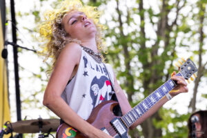Female guitarist playing a brown electric guitar on an outdoor stage at a festival venue.
