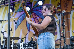Woman guitarist on stage, playing a wooden bass guitar with a colorful backdrop and drum set in the background.