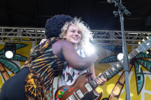 Two musicians share a hug on stage, smiling, with a brown electric guitar in the foreground against a colorful backdrop.