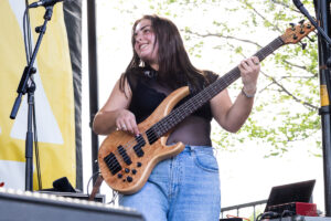 Female musician playing a natural wood bass guitar on an outdoor stage, smiling while performing in a black top and jeans.