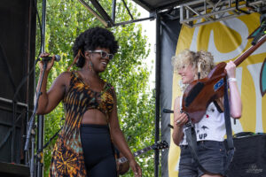 Two female musicians performing on an outdoor stage: one sings into a microphone while the other plays a violin and smiles nearby.
