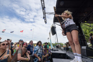 Female guitarist on stage with guitar raised, performing to cheering crowd at an outdoor festival under a cloudy sky.