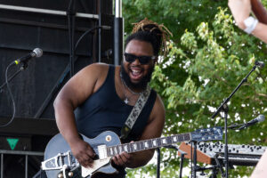 Muscular guitarist wearing sunglasses and a black tank top plays a gray LP-style guitar on an outdoor stage, with trees in the background and a keyboard setup nearby.