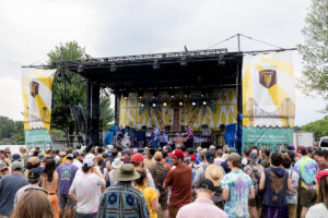 Outdoor concert stage with a band performing as a crowd watches in front of a festival setup