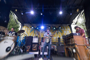 Live outdoor rock band on stage: guitarist center, drummer left, keyboardist right, with a yellow palm-tree backdrop.