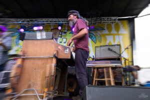Male musician with long hair and beard, wearing a maroon shirt and cap, playing a wooden keyboard on a colorful festival stage backdrop with purple lights.