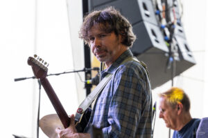 Guitarist in a plaid shirt playing an electric guitar on stage, with another musician in the background.