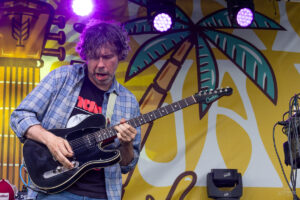 Male guitarist wearing a blue plaid shirt plays a black electric guitar on a colorful stage backdrop.