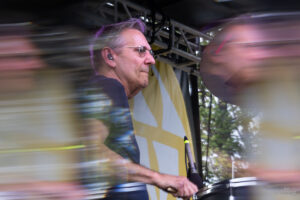 A middle-aged man wearing glasses drums on a stage, with motion blur from movement and a yellow banner in the background behind him.