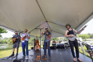 Band performing on an outdoor stage under a large tent by a lake, with guitarists, a violinist, and a banjo player.