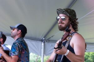 Bearded guitarist wearing a bandana hat and sunglasses, playing an acoustic guitar under a tent at an outdoor festival.