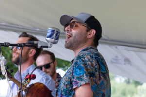 Male singer wearing a gray cap and sunglasses, singing into a retro microphone on a stage with band members in the background.
