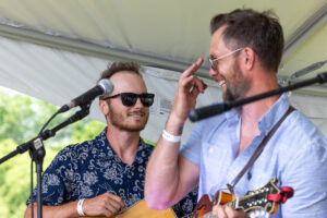 Two musicians on a sunny stage, one wearing sunglasses and a patterned shirt, smiling while the other strums a guitar and gestures playfully at the microphone.