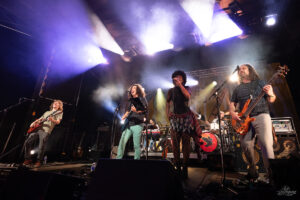 A live rock band on stage performing under colorful lights, with guitarists, a vocalist, and a drummer in the background.