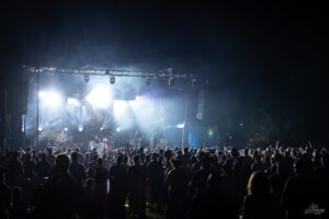 Nighttime outdoor concert with a band on stage and a large crowd watching the performance under bright stage lights.